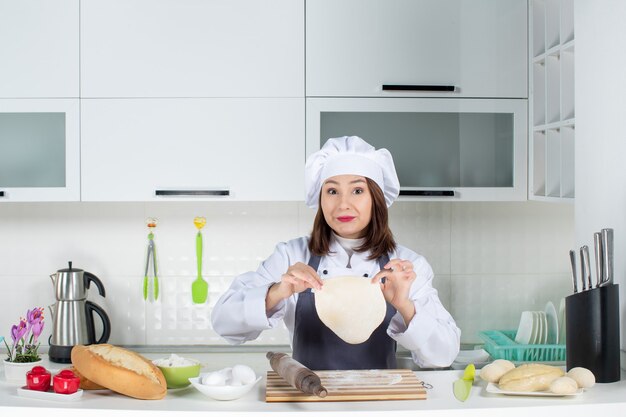 Cocinero de comis femenino feliz y positivo en uniforme de pie detrás de la mesa preparando pasteles en la cocina blanca