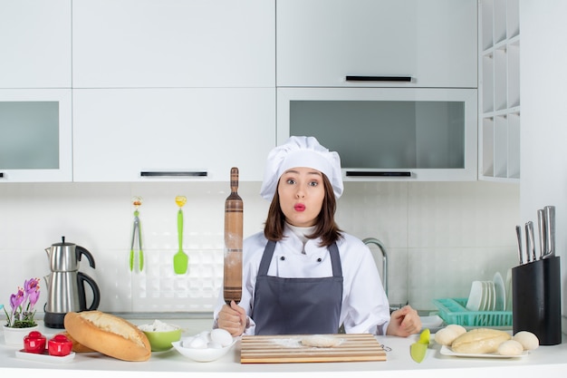 Cocinero de comis femenino sorprendido en uniforme de pie detrás de la mesa preparando pasteles en la cocina blanca