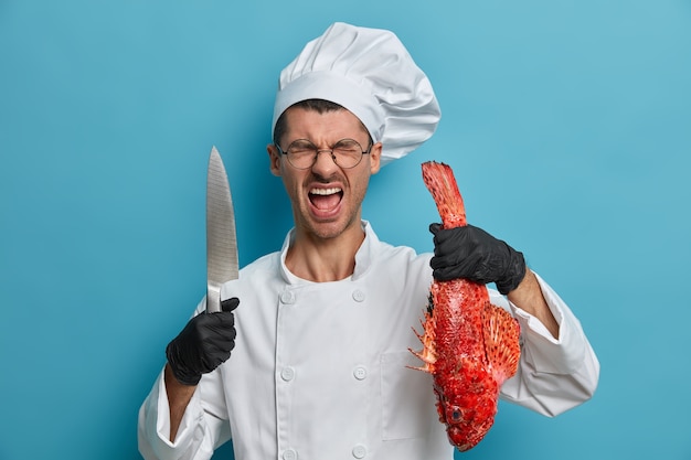 Cocinero francés profesional enojado pasar mucho tiempo en la cocina, viste uniforme, guantes de goma negros, posa con pescado y cuchillo