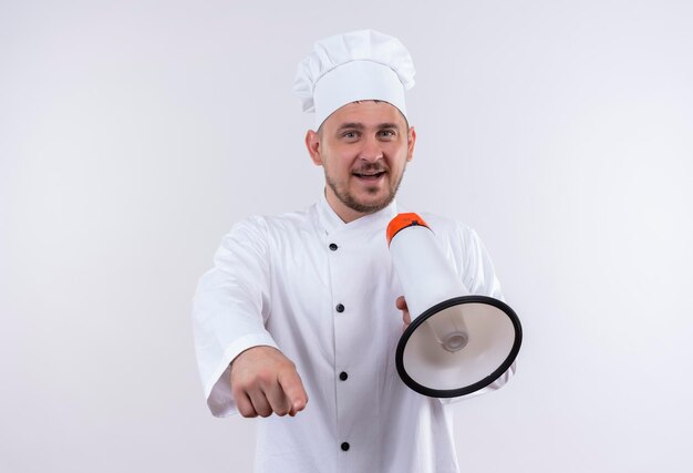 Cocinero guapo joven alegre en uniforme de chef sosteniendo el altavoz y apuntando aislado en la pared blanca