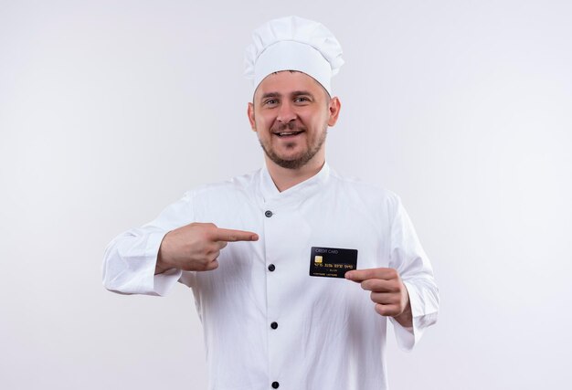Foto gratuita cocinero guapo joven alegre en uniforme de chef sosteniendo y apuntando a la tarjeta de crédito aislada en la pared blanca