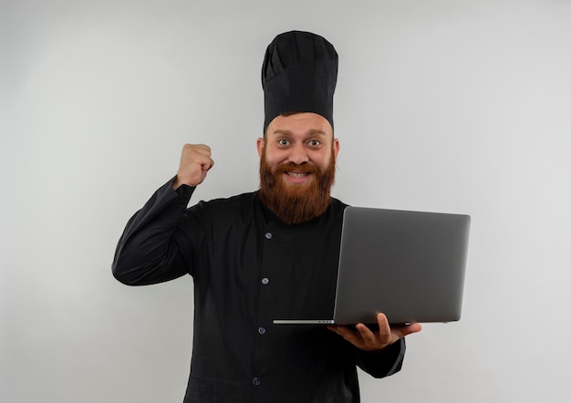 Cocinero guapo joven alegre en uniforme de chef sosteniendo el portátil y levantando el puño aislado en la pared blanca