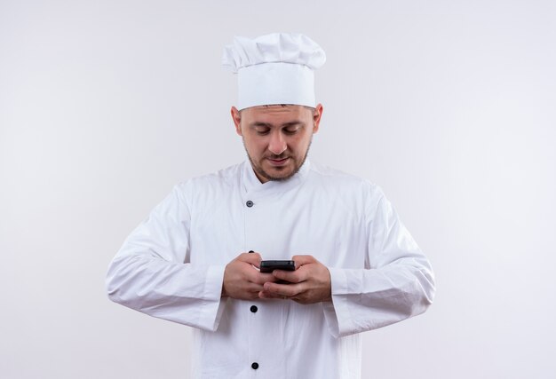 Cocinero guapo joven complacido en uniforme de chef sosteniendo y usando teléfono móvil aislado en el espacio en blanco