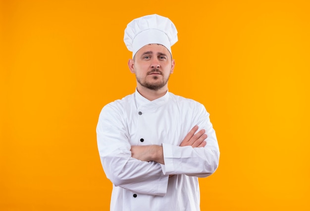 Cocinero guapo joven confiado en uniforme de chef de pie con postura cerrada aislada en la pared naranja