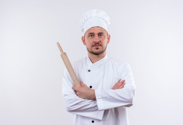 Cocinero guapo joven confiado en uniforme de chef de pie con postura cerrada y sosteniendo el rodillo aislado en la pared blanca