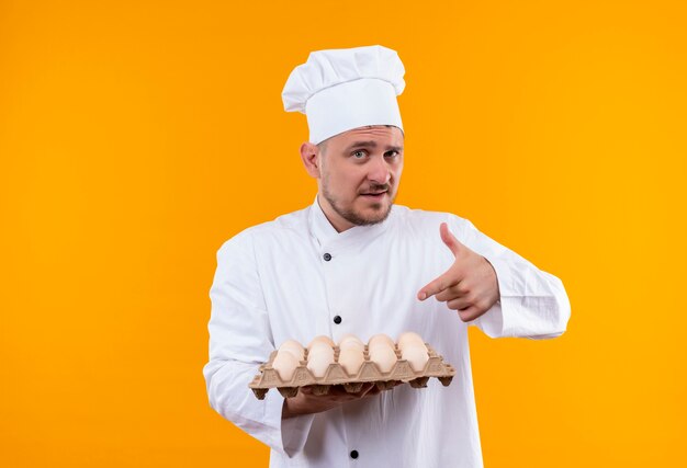 Cocinero guapo joven confiado en uniforme de chef sosteniendo y apuntando al cartón de huevos aislado en la pared naranja