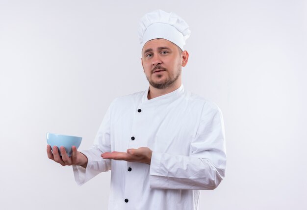 Cocinero guapo joven confiado en uniforme de chef sosteniendo y apuntando al recipiente aislado en la pared blanca