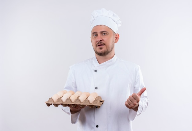 Cocinero guapo joven confiado en uniforme de chef sosteniendo cartón de huevos y mostrando el pulgar hacia arriba aislado en la pared blanca