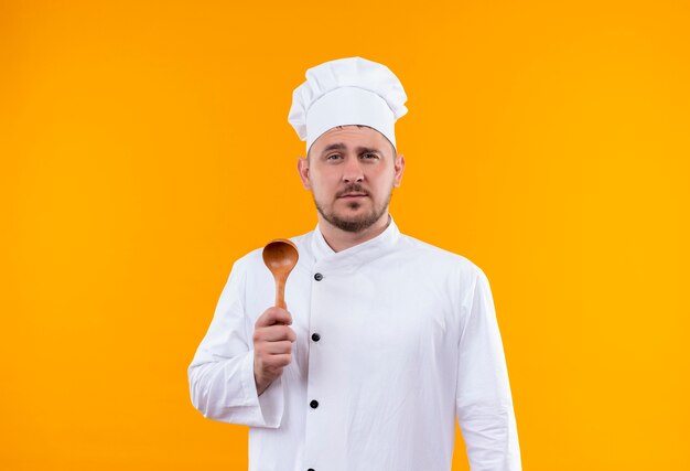 Cocinero guapo joven confiado en uniforme de chef sosteniendo una cuchara de madera aislada en la pared naranja