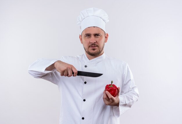 Cocinero guapo joven confiado en uniforme de chef sosteniendo pimienta y apuntando con un cuchillo aislado en la pared blanca