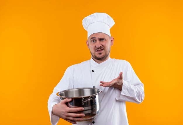 Cocinero guapo joven confundido en uniforme de chef sosteniendo la caldera y mostrando la mano vacía en la pared naranja aislada