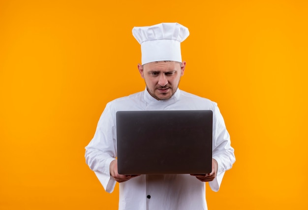 Cocinero guapo joven confuso en uniforme de chef sosteniendo y mirando portátil aislado en pared naranja