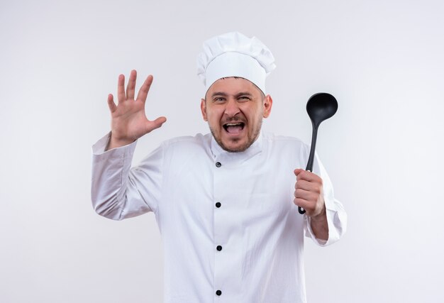 Cocinero guapo joven enojado en uniforme de chef sosteniendo la cuchara con la mano levantada en la pared blanca aislada