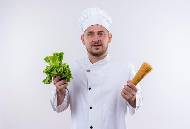 Cocinero guapo joven impresionado en uniforme de chef con lechuga y espaguetis aislado en la pared blanca