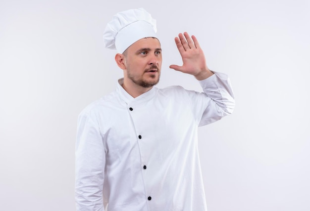 Cocinero guapo joven impresionado en uniforme de chef mirando al lado con la mano levantada aislado en la pared blanca