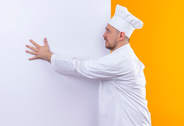 Cocinero guapo joven impresionado en uniforme de chef de pie delante de la pared blanca poniendo la mano en él mirando al lado aislado en la pared naranja