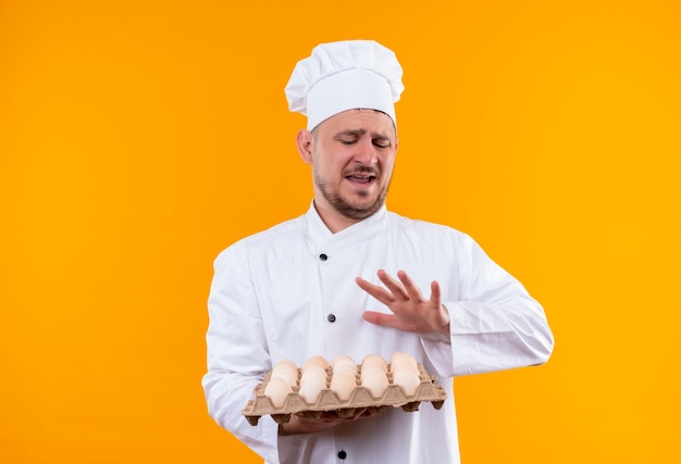 Cocinero guapo joven molesto en uniforme de chef sosteniendo cartón de huevos gesticulando no aislado en la pared naranja