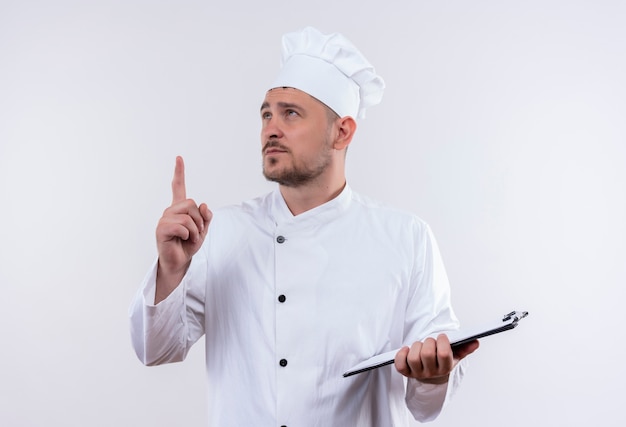Cocinero guapo joven pensativo en uniforme de chef sosteniendo portapapeles mirando y apuntando hacia arriba en el espacio en blanco aislado