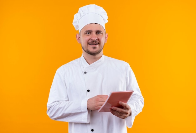 Cocinero guapo joven sonriente en uniforme de chef sosteniendo bloc de notas aislado en espacio naranja