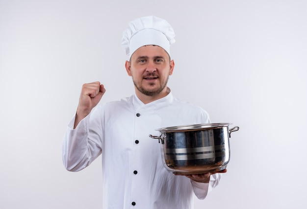 Cocinero guapo joven sonriente en uniforme de chef sosteniendo la olla y levantando el puño aislado en el espacio en blanco