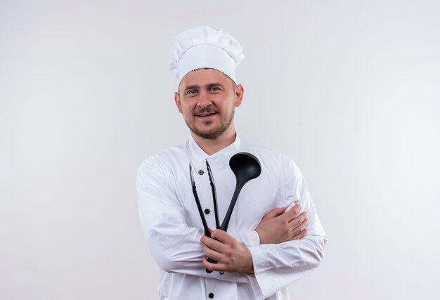Cocinero guapo joven sonriente en uniforme de chef sosteniendo pinzas de cuchara y parrilla aislado en el espacio en blanco