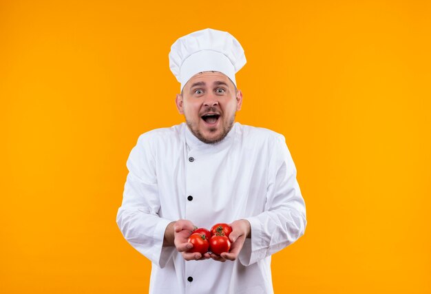 Cocinero guapo joven sorprendido en uniforme de chef sosteniendo tomates aislados en el espacio naranja