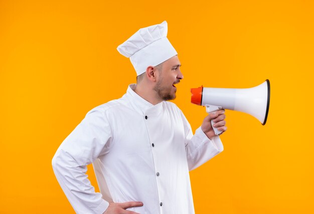 Cocinero guapo joven en uniforme de chef hablando por altavoz aislado en espacio naranja