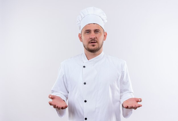 Cocinero guapo joven en uniforme de chef mostrando las manos vacías mirando aislado en el espacio en blanco