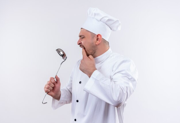 Cocinero guapo joven en uniforme de chef sosteniendo la cuchara mirándolo y poniendo el dedo en la boca en un espacio en blanco aislado