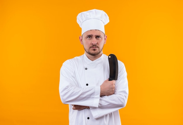 Cocinero guapo joven en uniforme de chef sosteniendo cuchillo mirando aislado en el espacio naranja