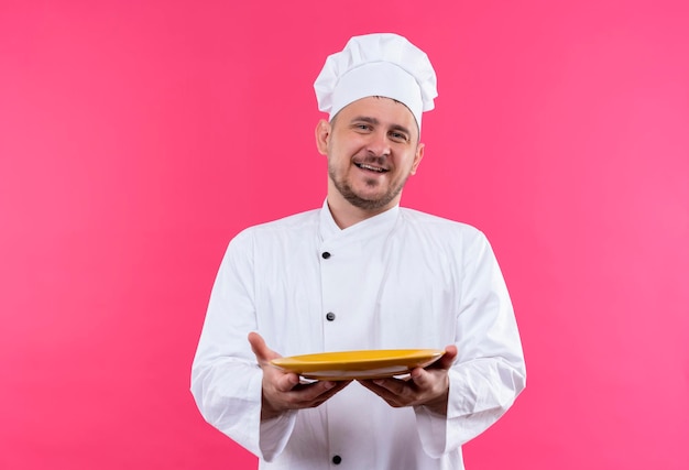 Cocinero hermoso joven alegre en uniforme del cocinero que se extiende el plato vacío aislado en la pared rosada