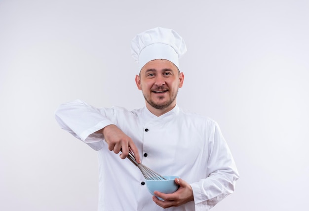 Cocinero hermoso joven alegre en uniforme del cocinero que sostiene el batidor y el cuenco en la pared blanca aislada