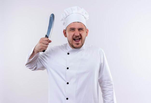Cocinero hermoso joven alegre en uniforme del cocinero que sostiene el cuchillo en la pared blanca aislada