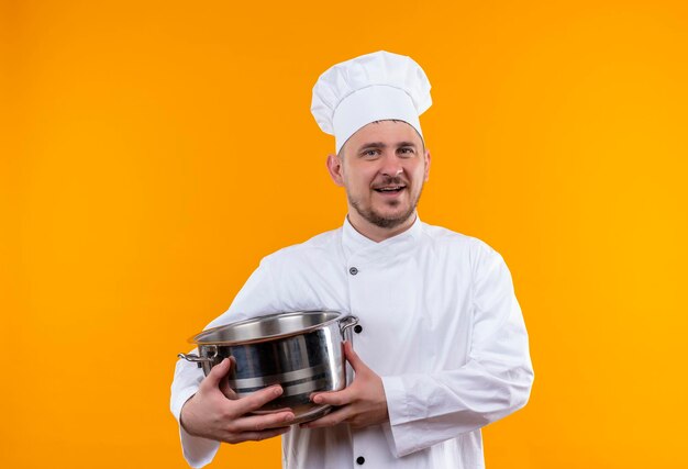 Cocinero hermoso joven alegre en uniforme del cocinero que sostiene la olla aislada en la pared naranja