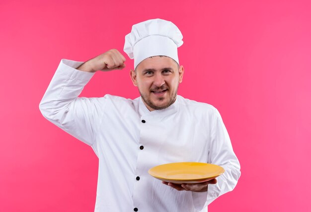 Cocinero hermoso joven alegre en el uniforme del cocinero que sostiene la placa que gesticula fuerte aislado en la pared rosada