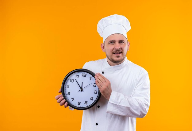 Cocinero hermoso joven alegre en uniforme del cocinero que sostiene el reloj aislado en la pared naranja