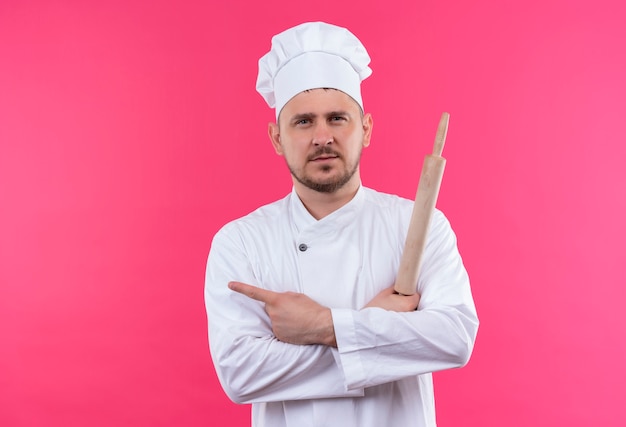 Cocinero hermoso joven confiado en uniforme del cocinero que sostiene el rodillo apuntando al lado aislado en la pared rosada