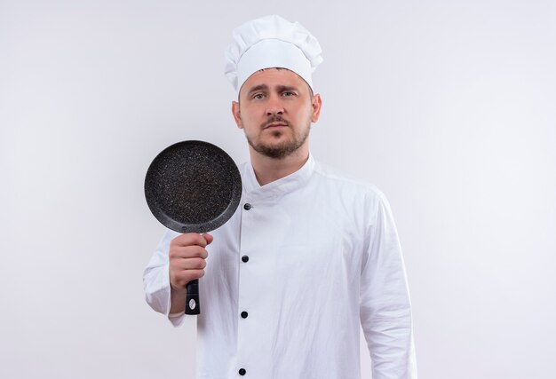 Cocinero hermoso joven confiado en uniforme del cocinero que sostiene la sartén en la pared blanca aislada