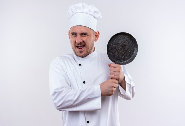 Cocinero hermoso joven confiado en uniforme del cocinero que sostiene la sartén en la pared blanca aislada