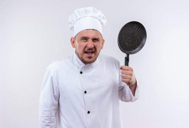 Cocinero hermoso joven enojado en uniforme del cocinero que sostiene la sartén aislada en la pared blanca