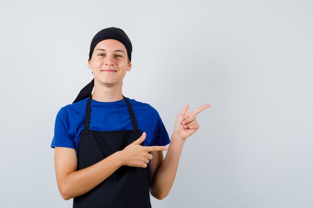 Cocinero joven en camiseta, delantal apuntando a la esquina superior derecha y mirando feliz, vista frontal.