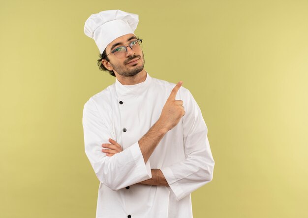 Cocinero joven complacido vistiendo uniforme de chef y gafas apunta al lado