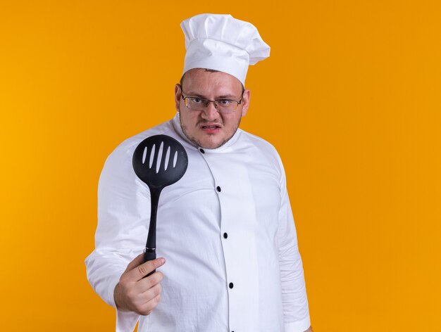 Cocinero macho adulto agresivo vistiendo uniforme de chef y gafas mirando al frente estirando una cuchara ranurada hacia el frente aislado en la pared naranja con espacio de copia