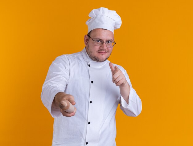 Cocinero macho adulto complacido con uniforme de chef y gafas mirando y apuntando al frente con el dedo y el rodillo aislado en la pared naranja con espacio de copia