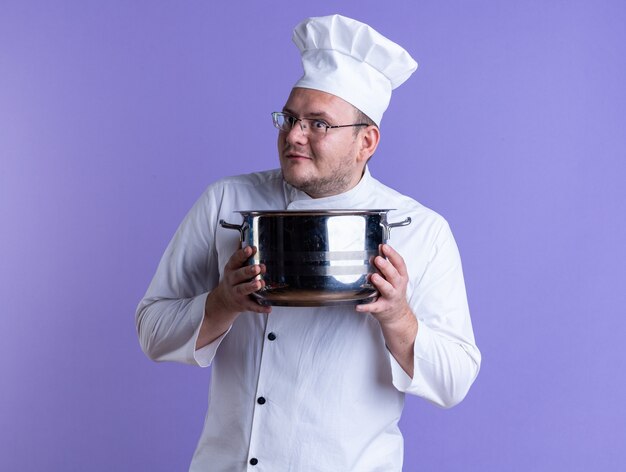Cocinero macho adulto impresionado vistiendo uniforme de chef y gafas sosteniendo la olla mirando al frente aislado en la pared púrpura