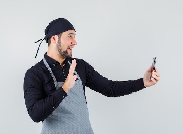 Cocinero masculino agitando la mano mientras toma selfie en camisa, delantal y mirando alegre, vista frontal.
