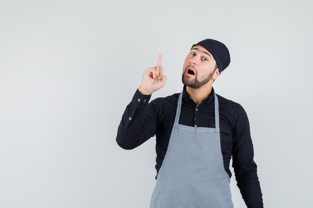 Cocinero masculino apuntando hacia arriba en camisa, delantal y mirando curioso. vista frontal.