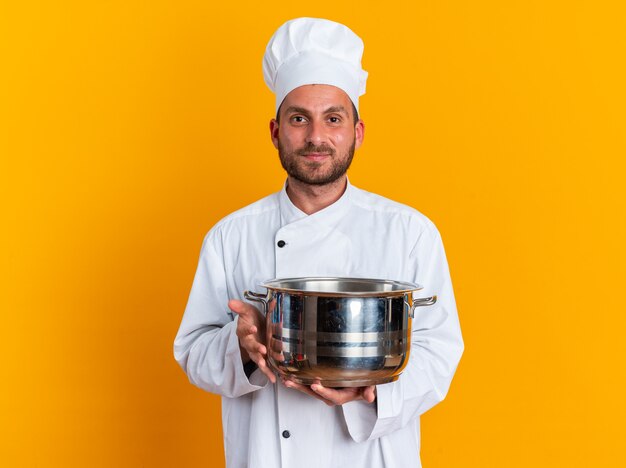 Cocinero masculino caucásico joven complacido en uniforme del cocinero y la tapa que sostiene la olla