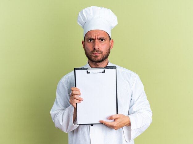 Cocinero masculino caucásico joven estricto en uniforme del cocinero y la tapa que muestra el portapapeles mirando a la cámara aislada en la pared verde oliva