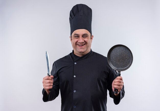 Cocinero masculino de mediana edad sonriente en uniforme del cocinero que sostiene la sartén y la cuchilla con espacio de copia